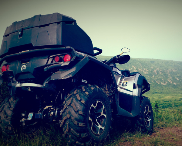 Schwarzes ATV mit robustem Koffer steht auf Wiese vor Hügelkulisse bei bewölktem Himmel.