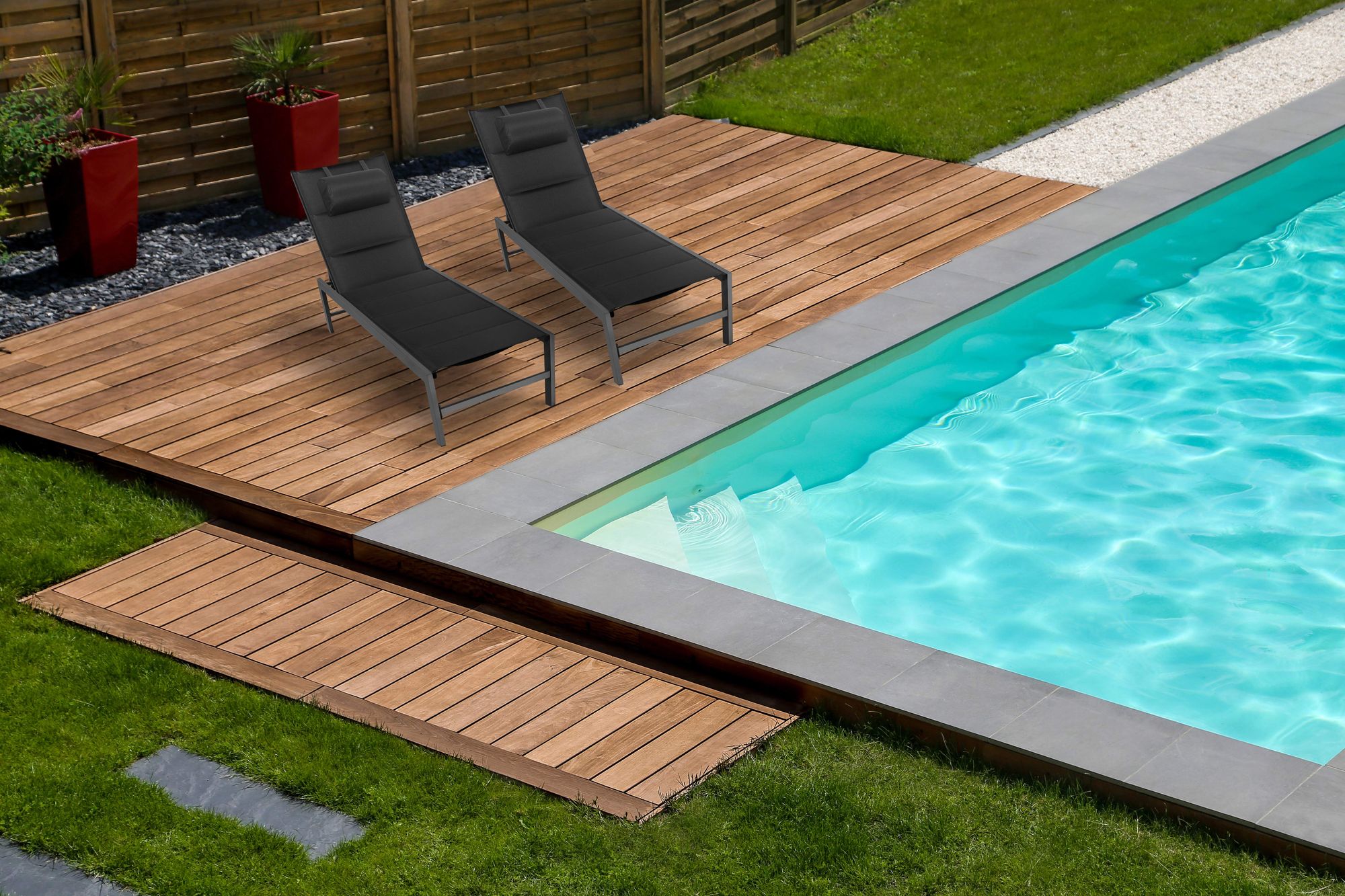 Pool mit Holzdeck und zwei schwarzen Sonnenliegen im gepflegten Garten bei Tageslicht.