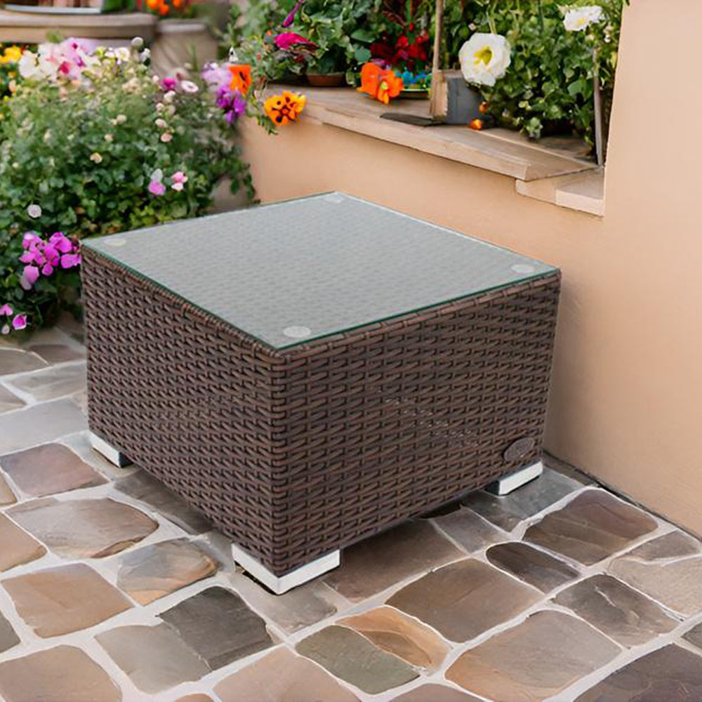 Brauner Rattan-Beistelltisch mit Glasplatte auf Steinboden, umgeben von Blumen und Pflanzen.