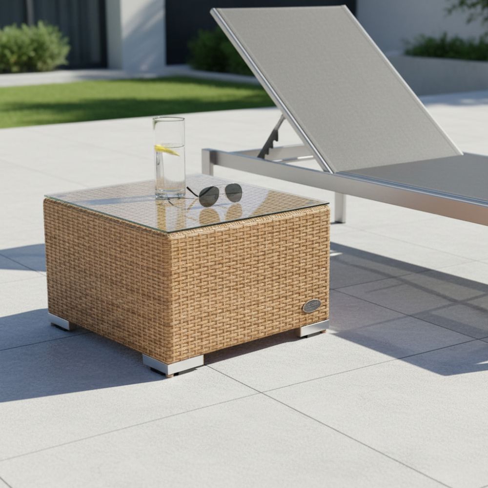 Quadratischer Beistelltisch aus naturfarbenem Polyrattan mit Glasplatte auf einer Terrasse neben einer Sonnenliege. Auf der Tischplatte stehen ein Getränk und eine Sonnenbrille.