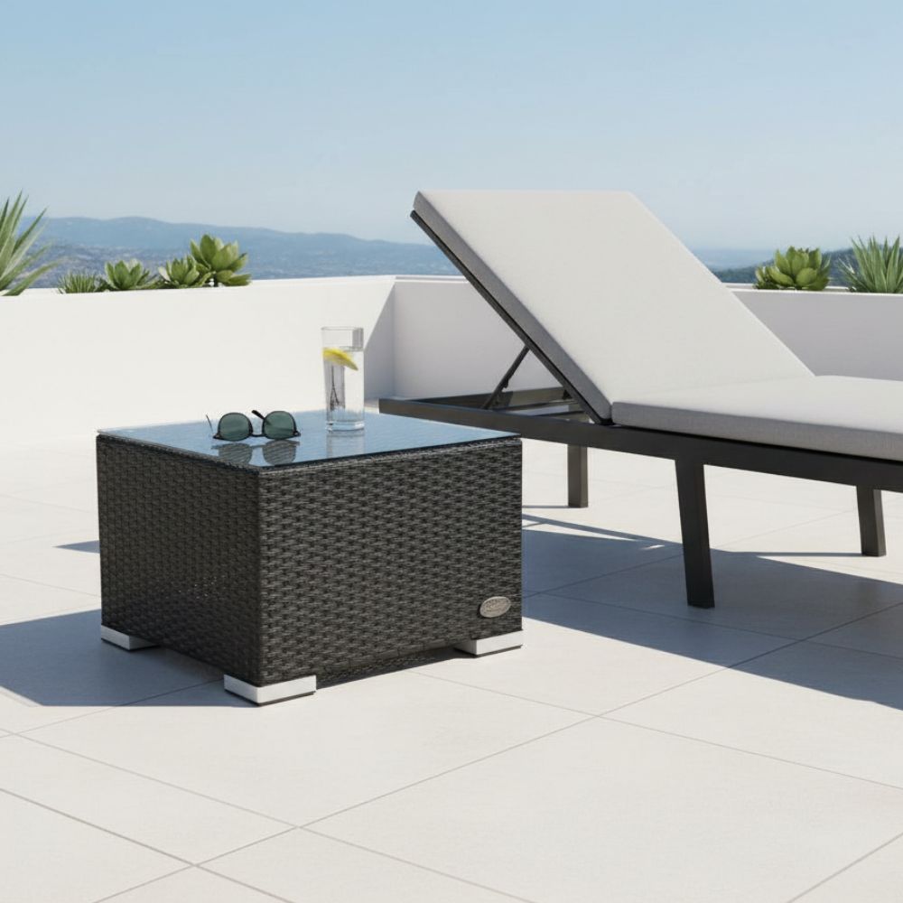 Schwarzer Polyrattan-Beistelltisch mit Glasplatte neben einer Sonnenliege auf einer Terrasse. Auf der Tischplatte befinden sich ein Getränk und eine Sonnenbrille.