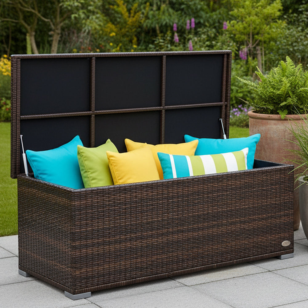 Garten-Auflagenbox aus braunem Polyrattan mit geöffnetem Deckel und bunten Kissen auf einer Terrasse im grünen Garten.