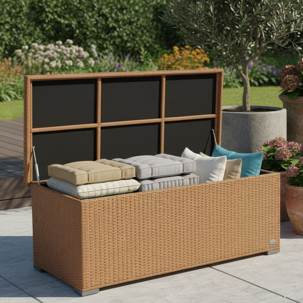 Naturbraune Rattanbox mit geöffnetem Deckel auf heller Terrasse, umgeben von grüner Bepflanzung.