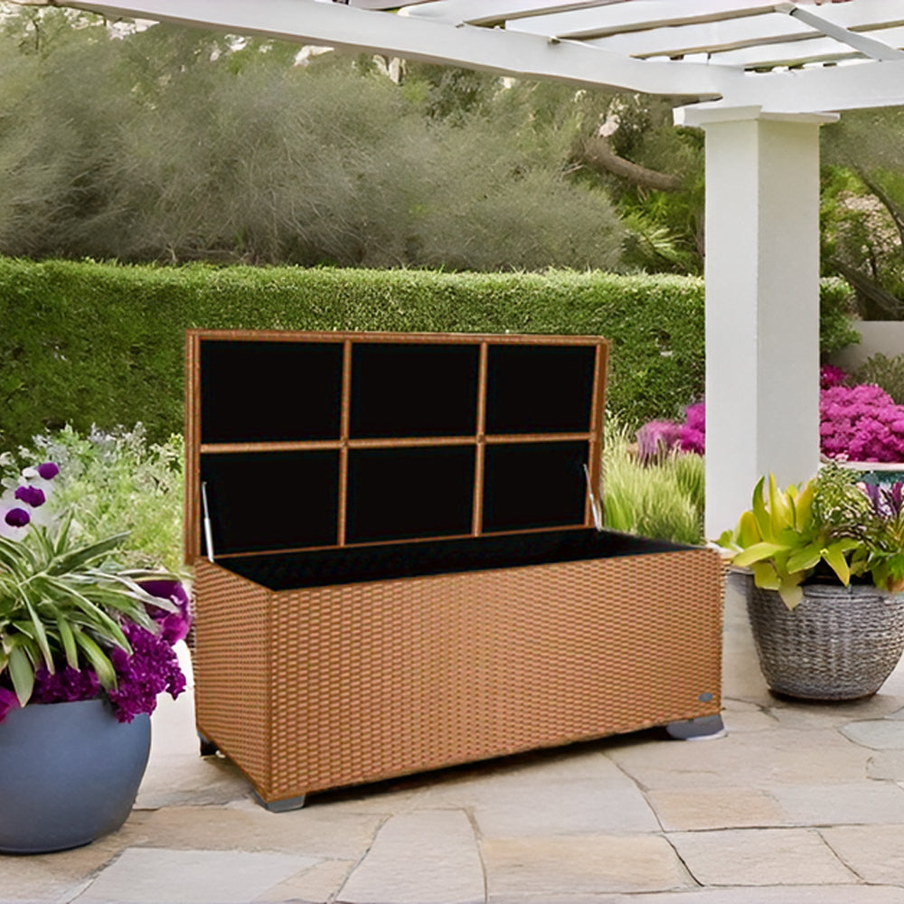 Geöffnete Aufbewahrungsbox in beiger Rattan-Optik auf einer Terrasse mit Steinbelag, daneben Pflanzen und im Hintergrund ein Garten.