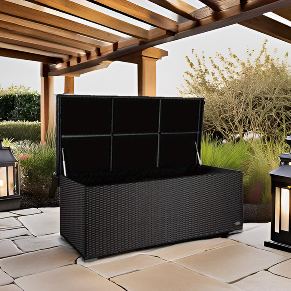 Geöffnete Aufbewahrungsbox in schwarzer Rattan-Optik auf einer Terrasse mit Steinbelag, daneben Laternen und im Hintergrund ein Garten.