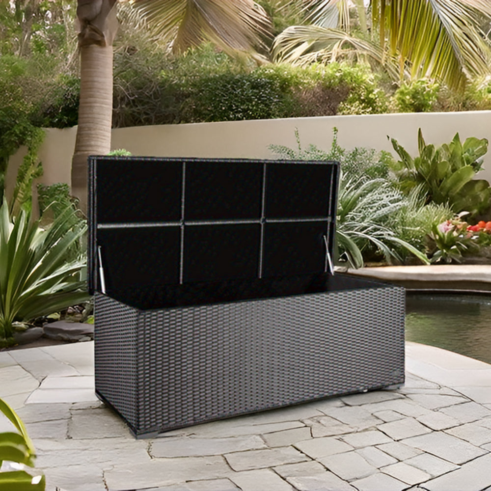 Geöffnete Aufbewahrungsbox in silber/grauer Rattan-Optik auf einer Terrasse mit Steinbelag, im Hintergrund ein Teich und Palmen.