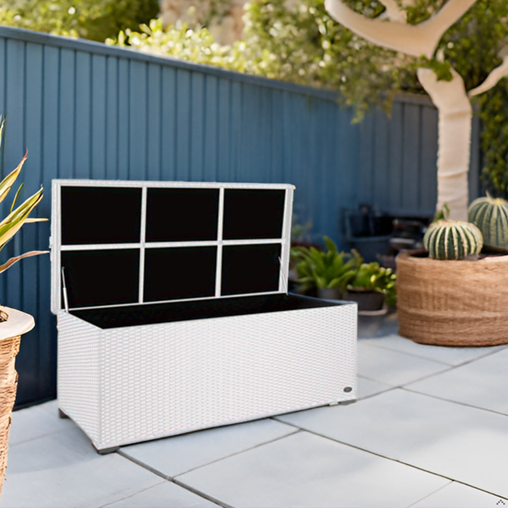 Geöffnete Aufbewahrungsbox in weißer Rattan-Optik auf einer Terrasse mit Steinbelag, daneben Pflanzen in Kübeln und ein blauer Zaun aus Holz.