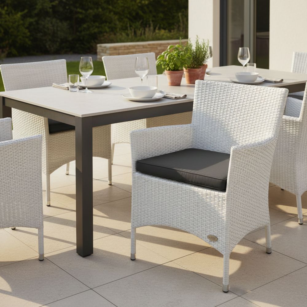 Weißer Polyrattan-Gartenstuhl „Trento“ mit anthrazitfarbenem Sitzkissen an modernem Esstisch auf sonniger Terrasse. Wetterfester Outdoor-Stuhl mit Armlehnen für Garten, Balkon und Terrasse.