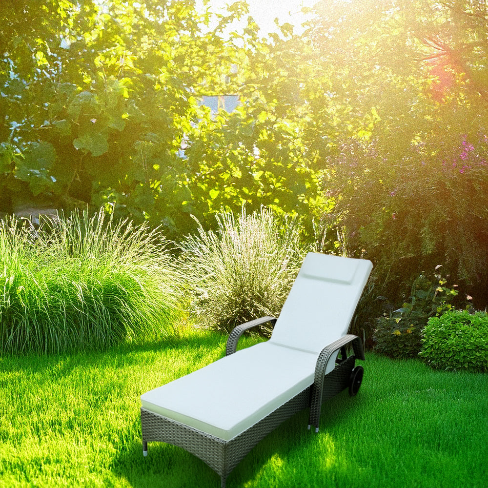 Grauer Rattan-Liegestuhl mit Polster steht in einem Garten zwischen grünen Pflanzen.