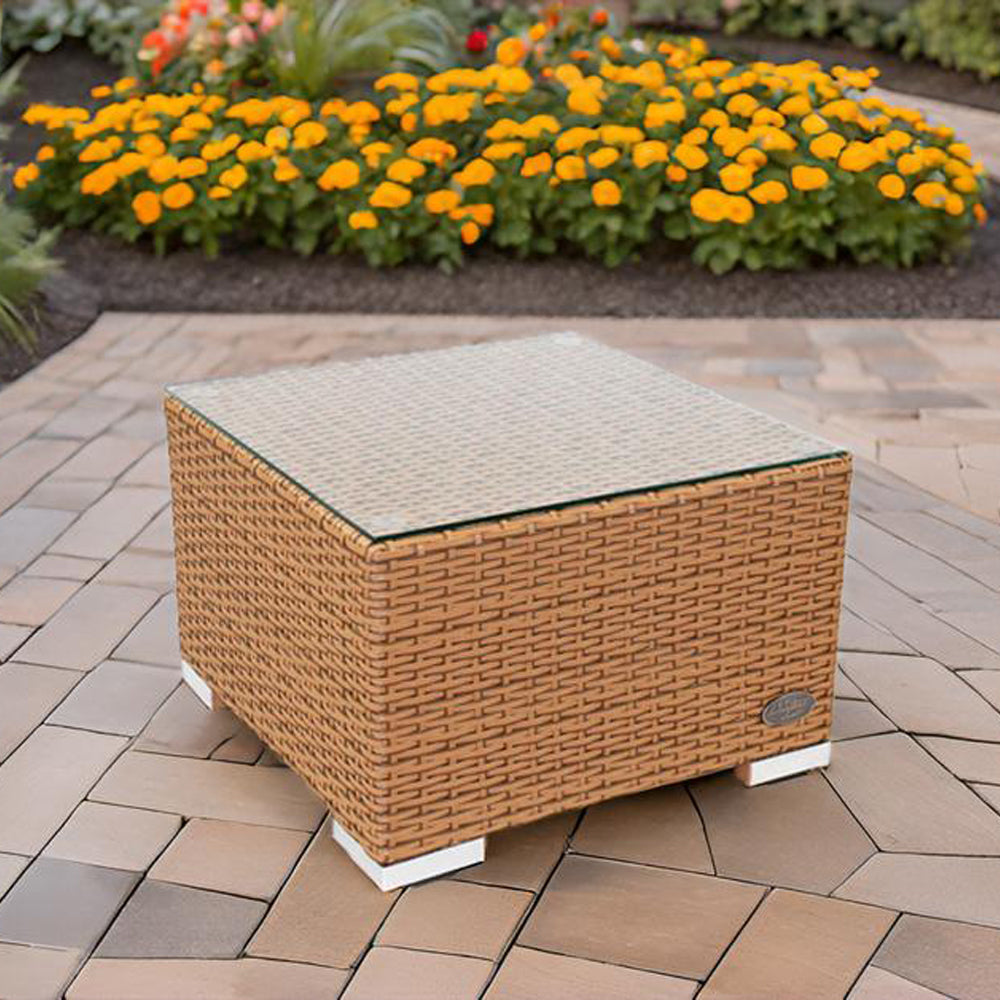 Naturfarbener Rattantisch mit Glasplatte von RS Trade, platziert auf Steinboden vor Blumenbeet.